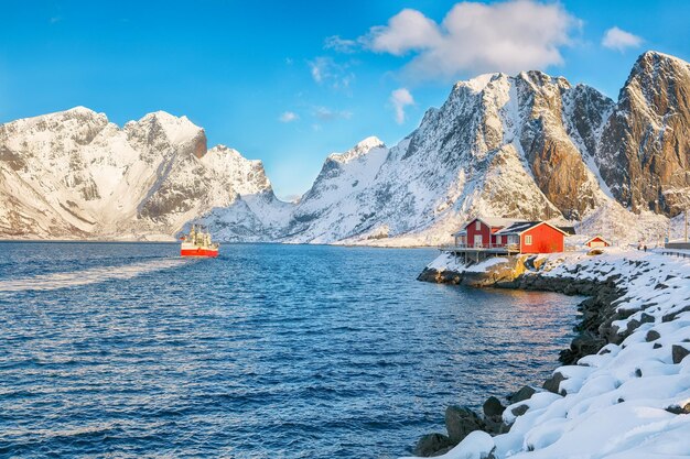 Traditional norwegian red wooden houses on the shore of reinefjorden and fishing ships with snowy mountaines in background