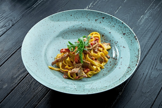 クリームソース、牛肉とトマト、ルッコラを添えた伝統的なパスタスパゲッティは、黒い木製の背景に青いプレートで提供されます。イタリア料理