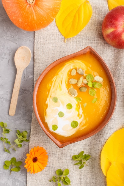 Traditional pumpkin cream soup with seeds in clay bowl on a gray concrete background with linen textile. top view