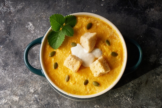 Traditional pumpkin soup puree with crackers, cream and seeds in a bowl, on a dark background