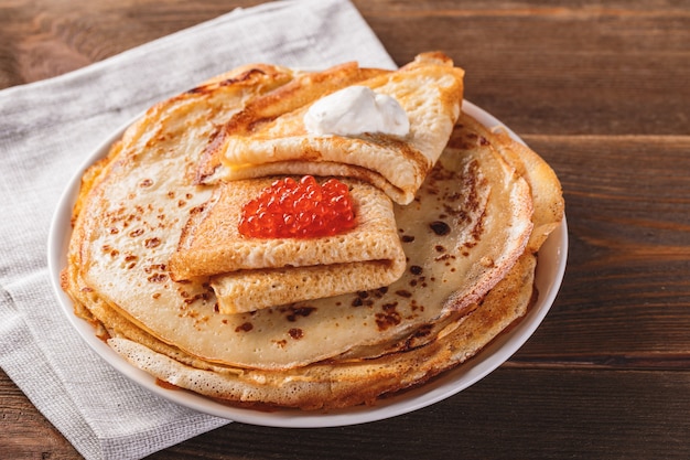 Traditional Russian Crepes Blini stacked in a plate with red caviar, fresh sour creamon dark wooden table. Maslenitsa traditional Russian festival meal. Russian food, russian kitchen