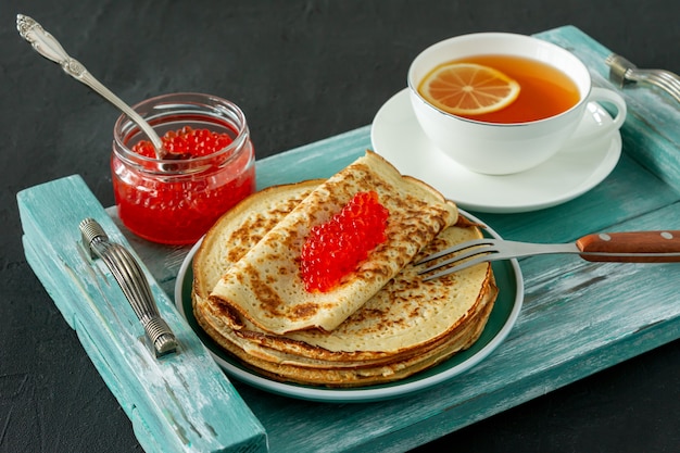 Traditional Russian Crepes Blini stacked in a plate with red caviar and hot lemon tea on wooden background. Maslenitsa traditional Russian festival meal. Russian food, russian kitchen.