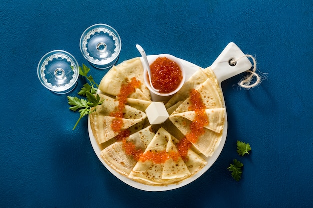 Traditional Russian pancakes with red caviar and butter on a blue table background and two glasses of vodka