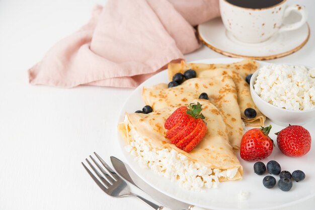 Traditional Russian thin pancakes with cottage cheese and berries in a plate on a white, selective focus, copy space