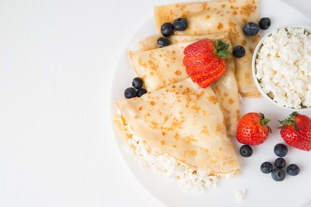 Traditional Russian thin pancakes with cottage cheese and berries in a plate on a white, top view, copy space