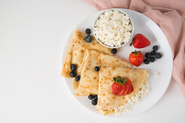 Frittelle sottili russe tradizionali con ricotta e frutti di bosco in un piatto su un bianco, vista dall'alto, copia dello spazio