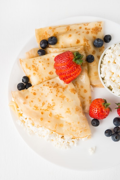 Traditional Russian thin pancakes with cottage cheese and berries in a plate on a white, top view