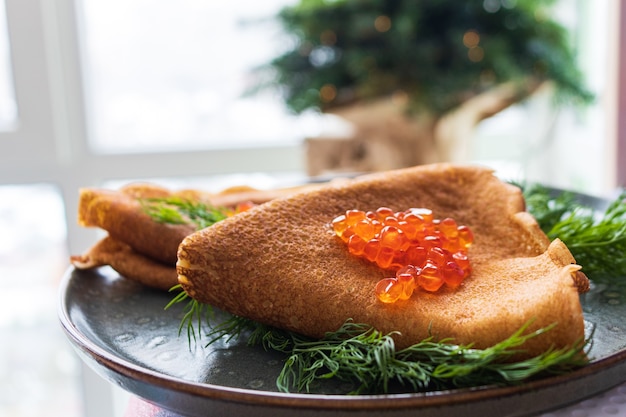 Traditional Russian thin pancakes with red caviar close-up with dill on the background of a Christmas tree with bokeh effect, blurred background