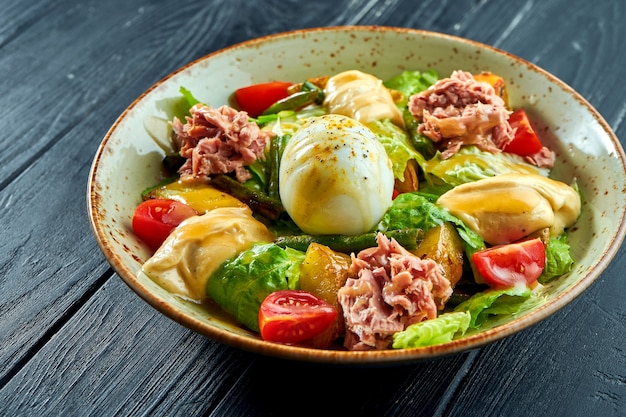 Traditional salad of French cuisine - nicoise with tuna, asparagus, tomatoes, potatoes and poached egg served in a bowl on a black wooden surface