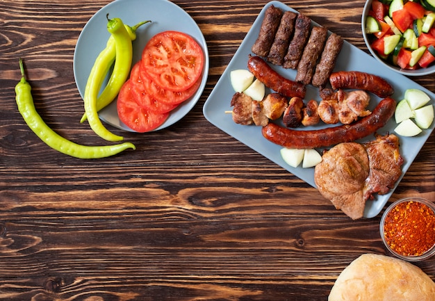 Traditional Serbian and Balkan grilled meat called mesano meso. Balkan barbeque (rostilj) served with Serbian salad, hot peppers, bread, tomato, onions, and paprika powder. Wooden background. Top view