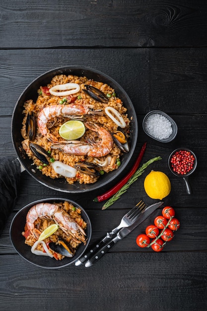 Традиционная испанская паэлья из морепродуктов в сковороде и миске с рисовым горохом, креветками, мидиями и кальмарами на черных деревянных досках, вид сверху