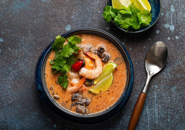 Traditional spicy Thai soup Tom Yum with shrimp and seafood in ceramic bowl on blue stone rustic background from above, classic dish of cuisine of Thailand