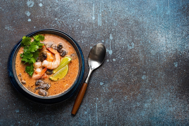 Traditional spicy Thai soup Tom Yum with shrimp and seafood in ceramic bowl on blue stone rustic background from above with space for text, classic dish of cuisine of Thailand