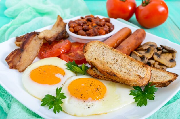 Traditioneel Engels ontbijt: spek, champignons, eieren, tomaten, worstjes, bonen, toast op een witte plaat op een lichte houten tafel. Engeland klassieke keuken.