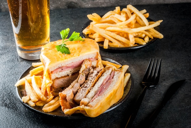 Traditioneel Portugees snackvoedsel. Broodje Francesinha van brood, kaas, varkensvlees, ham, worstjes, met tomatensaus en frites. Met een glas bier en aardappelen. Op zwarte tafel.