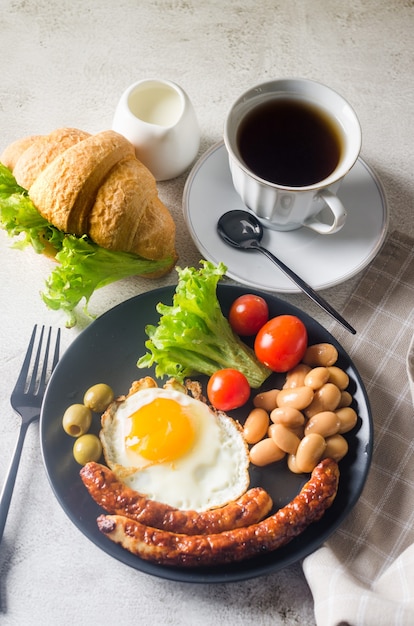 Traditioneel volledig Engels ontbijt met gebakken eieren, worstjes, bonen, tomaten op een bord, croissant en koffie op grijze achtergrond. Uitzicht van boven