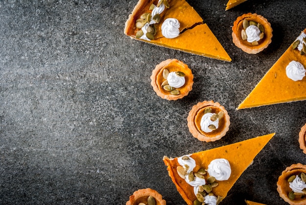 Traditionele herfstgerechten. Halloween, Thanksgiving. Set van pittige pompoentaart en pompoentaartjes met slagroom en pompoenpitten op zwarte stenen tafel. bovenaanzicht