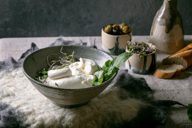 Traditionele italiaanse burrata geknoopte kaassalade in grijze keramische kom op tafel met vilten servet. Gesneden brood, olijven, groene spruiten rond. Gezond mediterraan diner.