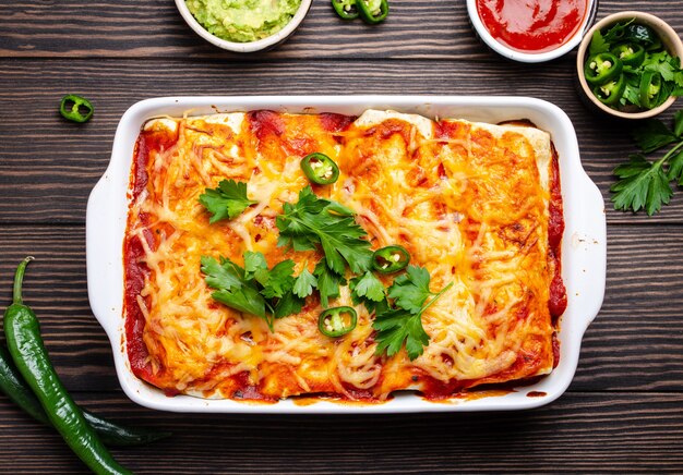 Traditionele Mexicaanse schotel enchiladas met vlees, rode chilisaus en kaas in witte braadpan schotel op rustieke houten achtergrond, geserveerd met guacamole en tomaat dips. Close-up, bovenaanzicht