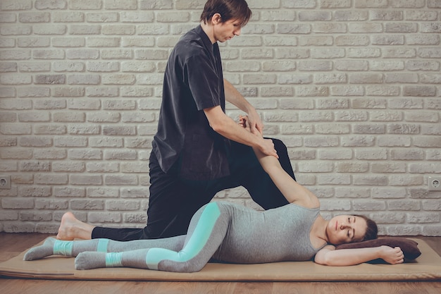 Traditionele Thaise Massage van zwangere vrouw liggend op de mat in yogastudio. Jonge witte masseur gekleed in een zwart uniform strekt haar hand uit. Bakstenen muur op de achtergrond. Gezondheid, ontspanning, spa-concept