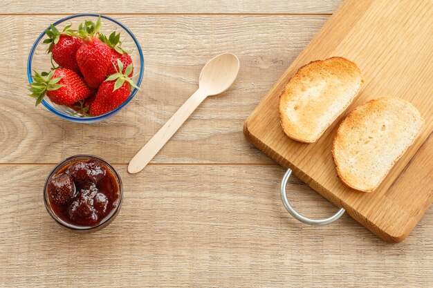 Traditionele zelfgemaakte aardbeienjam en verse bessen in glazen kommen, houten lepel en toast op snijplank op houten bureau. bovenaanzicht.