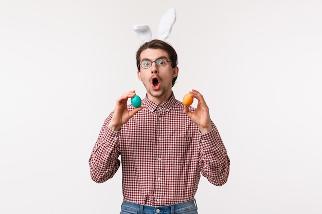 전통, 종교 휴일, 축 하 개념. 안경에 수염을 가진 재미있는 즐거운 젊은이, 귀여운 토끼 귀를 착용하고 부활절 날, 흰 벽에 게임을하는 두 개의 그려진 계란을 잡아