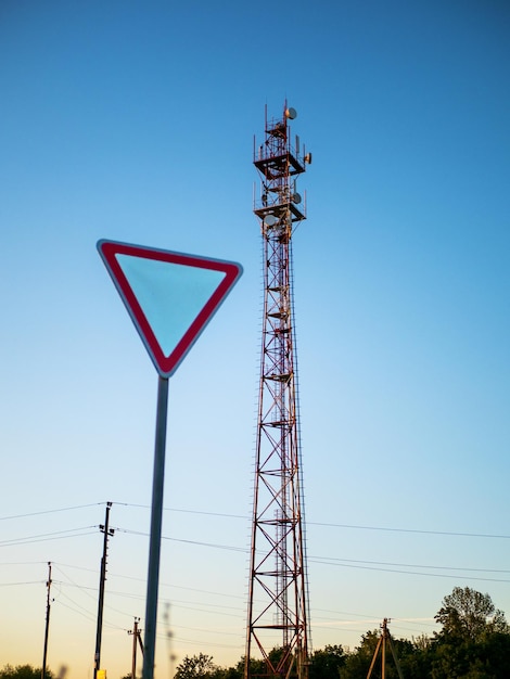 Traffic sign give way against the backdrop of a cellular repeater tower against a blue sunset sky