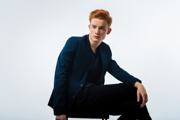 Train of thoughts. Good-looking stern dark-eyed red-haired young man wearing a black jacket and thinking and sitting on the chair