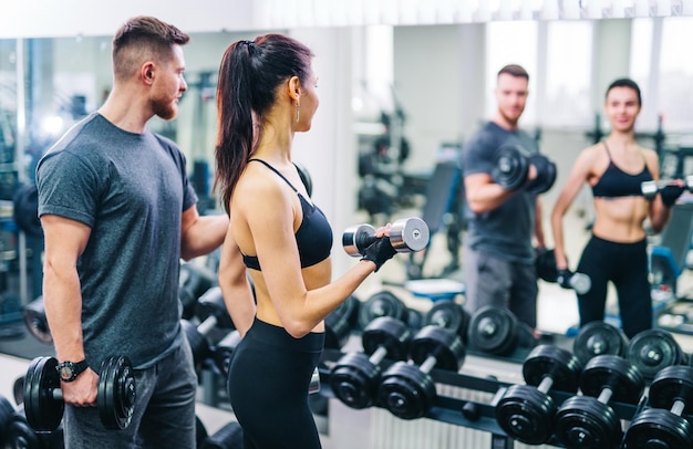 Foto allenatore e cliente che si guardano allo specchio mentre si allena in palestra con manubri giovane bruna con personal fitness trainer concetto di fitness primo piano messa a fuoco selettiva