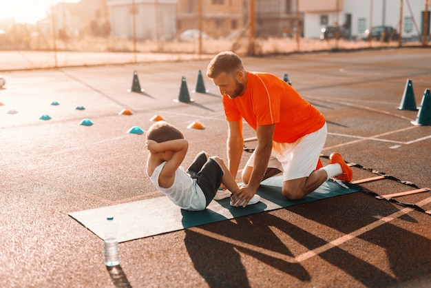 Trainer helping kid to do abs on the court in the morning.