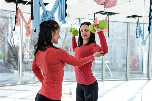 Тренировки подходят девушке вместе. Портрет двух здоровых молодых женщин, занимающихся фитнесом в ярком тренажерном зале