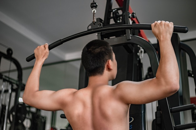 Training gym concept a male teenager using a gym equipment pulling his both muscular arms against the machine.