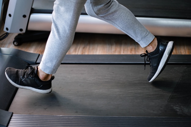 Концепция тренажерного зала мужской подросток в длинных штанах и черных кроссовках, идущих на беговой дорожке для кардиотренировки.