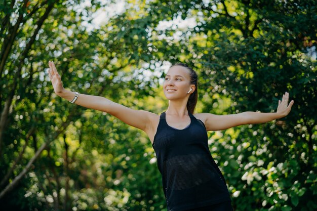 Foto concetto di allenamento all'aperto la donna europea sportiva soddisfatta sente che la libertà ti motiva per lo sport allunga le braccia lateralmente conduce uno stile di vita attivo indossa una maglietta nera posa contro alberi verdi all'esterno