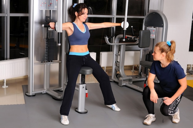 Training sportive brunette girl sitting on fitness machine and exercise arm muscles under the supervision of trainer