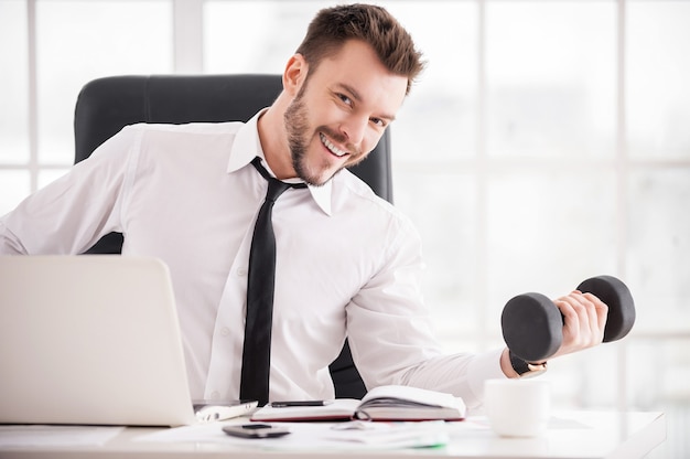 Training at the working place. Handsome young beard man in shirt and tie training with dumbbell and smiling at camera while sitting at his working place