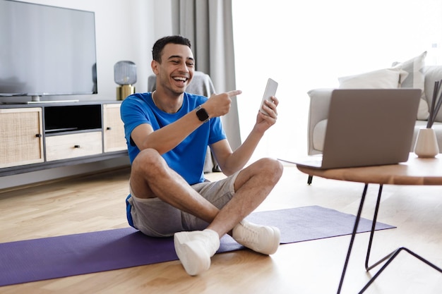 Trainingstoepassing Gelukkige mannelijke fitnessblogger die smartphone toont aan laptop-webcam die app voor training aanbeveelt