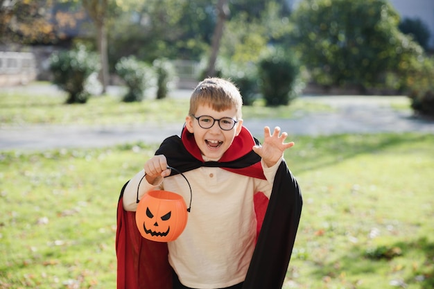 Trakteer of trick Buitenopname van kleine jongen in vampier-carnaval-outfit voor Halloween die in het park staat met pompoenmand in zijn hand