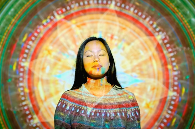 얼굴에 빛이 있고 눈이 닫힌 조용한 아시아 여성이 창의적인 장식으로 밝은 반 ⁇ 이는 만달라와 함께 벽 근처에 서서 스튜디오에서 명상하고 있습니다.