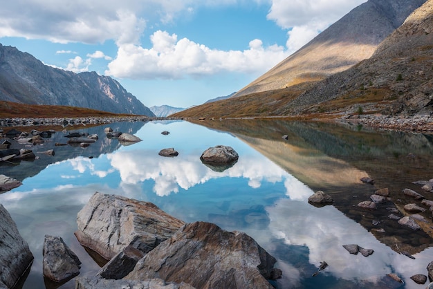 Tranquil autumn landscape with clouds reflection on smooth mirror surface of mountain lake in high hanging valley Meditative view from calm alpine lake to mountain vastness Stones in clear water