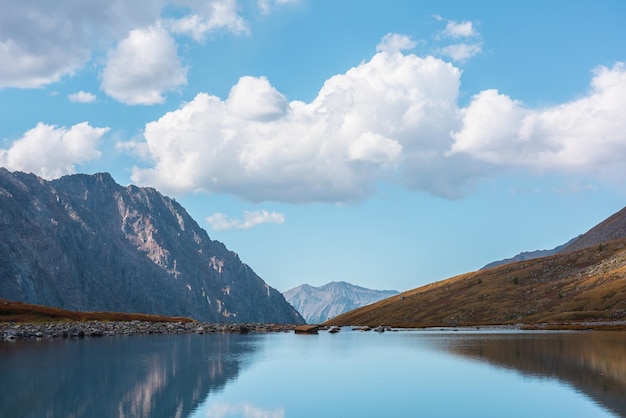青い空の雲の下、高く垂れ下がった谷の山の湖の滑らかな表面を持つ静かな秋の風景 静かな高山湖から山の広大さへの瞑想的な眺め 氷河湖の澄んだ水