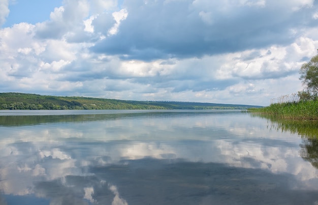 Tranquil landscape on the lake, with bright sky, white clouds and trees symmetrically reflected in clear blue water