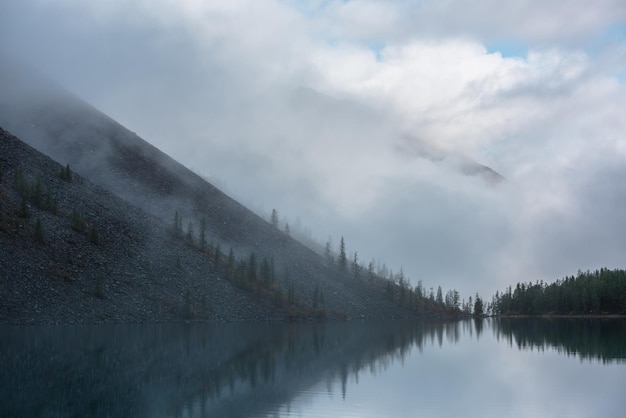 Tranquil meditative misty scenery of glacial lake with reflection of pointy fir tops and clouds at early morning Graphic EQ of spruce silhouettes on hill near calm alpine lake Mountain lake in fog