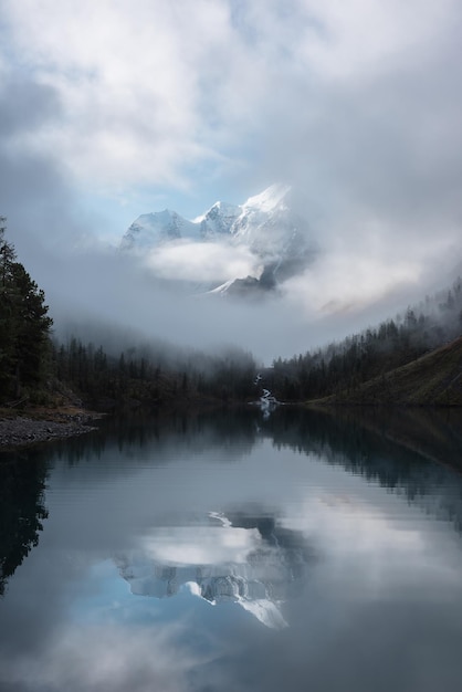 Tranquil scenery with snow castle in clouds Mountain creek flows from forest hills into glacial lake Snowy mountains in fog clearance small river and coniferous trees reflected in calm alpine lake