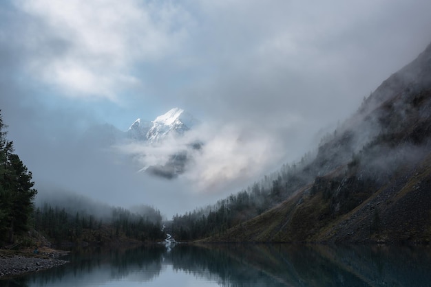 Photo tranquil scenery with snow castle in clouds mountain creek flows from forest hills into glacial lake snowy mountains in fog clearance small river and coniferous trees reflected in calm alpine lake