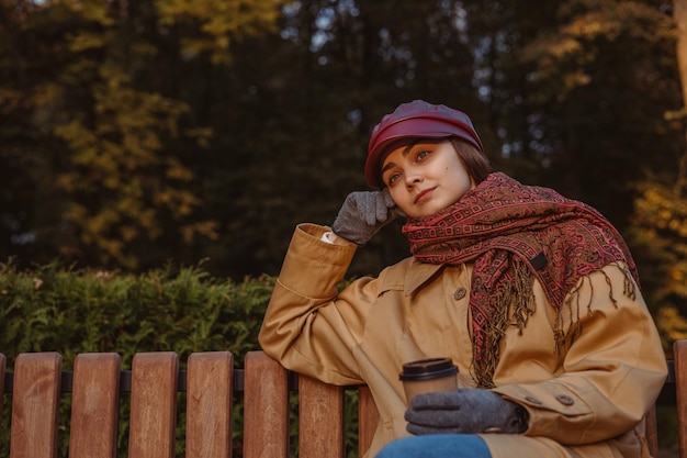 Tranquil woman in autumn wear with paper cup of coffee resting on bench in park at sunset