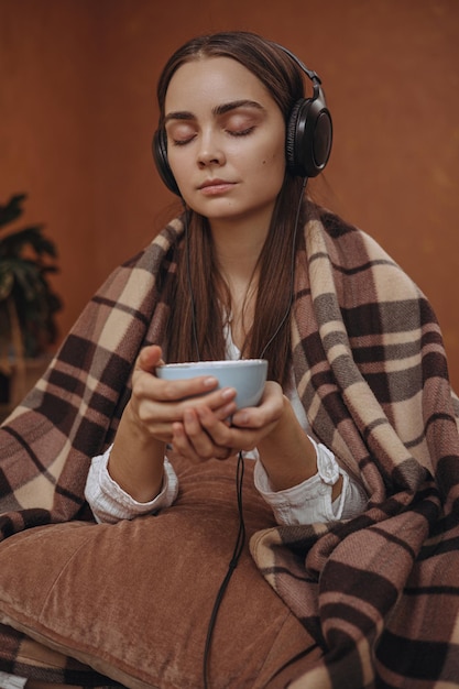 Tranquil woman in earphone listening to calm music and drinking tea while enjoying weekend at home