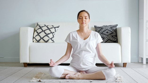 Tranquil young pregnant woman in white meditates sitting in lotus pose on floor rug near designer sofa in spacious room at home close-up