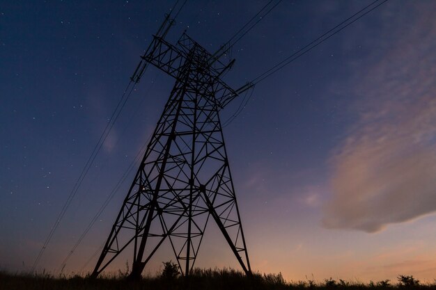 Transmission and long distance distribution of electricity concept. Angled view of high voltage tower with electric power lines stretching on dark blue starry sky scene.