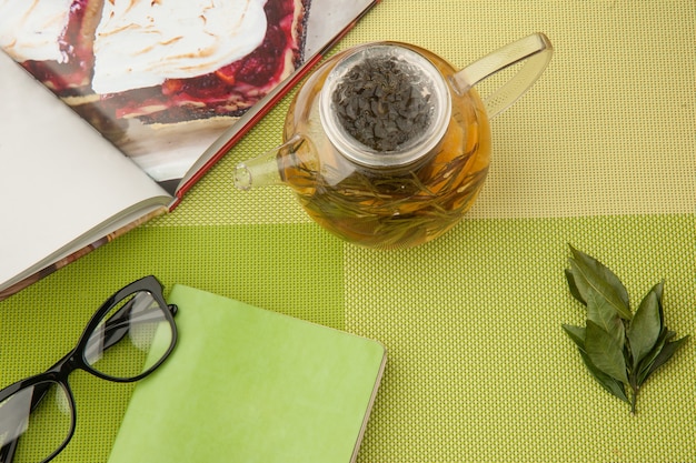 Transparante theepot met groene thee met boek en glazen op een groen tafelkleed